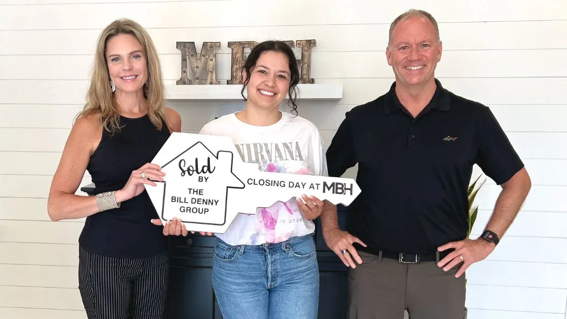 A homebuyer holding a Sold by The Bill Denny Group key sign on closing day at MBH Settlement Group with two Bill Denny Real Estate Group agents