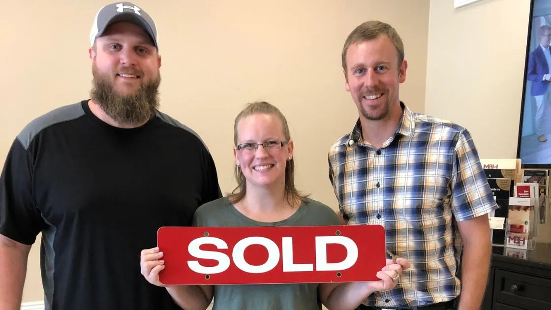 Happy homebuyers holding a SOLD sign with a Bill Denny Real Estate Group agent at the MBH Settlement Group office