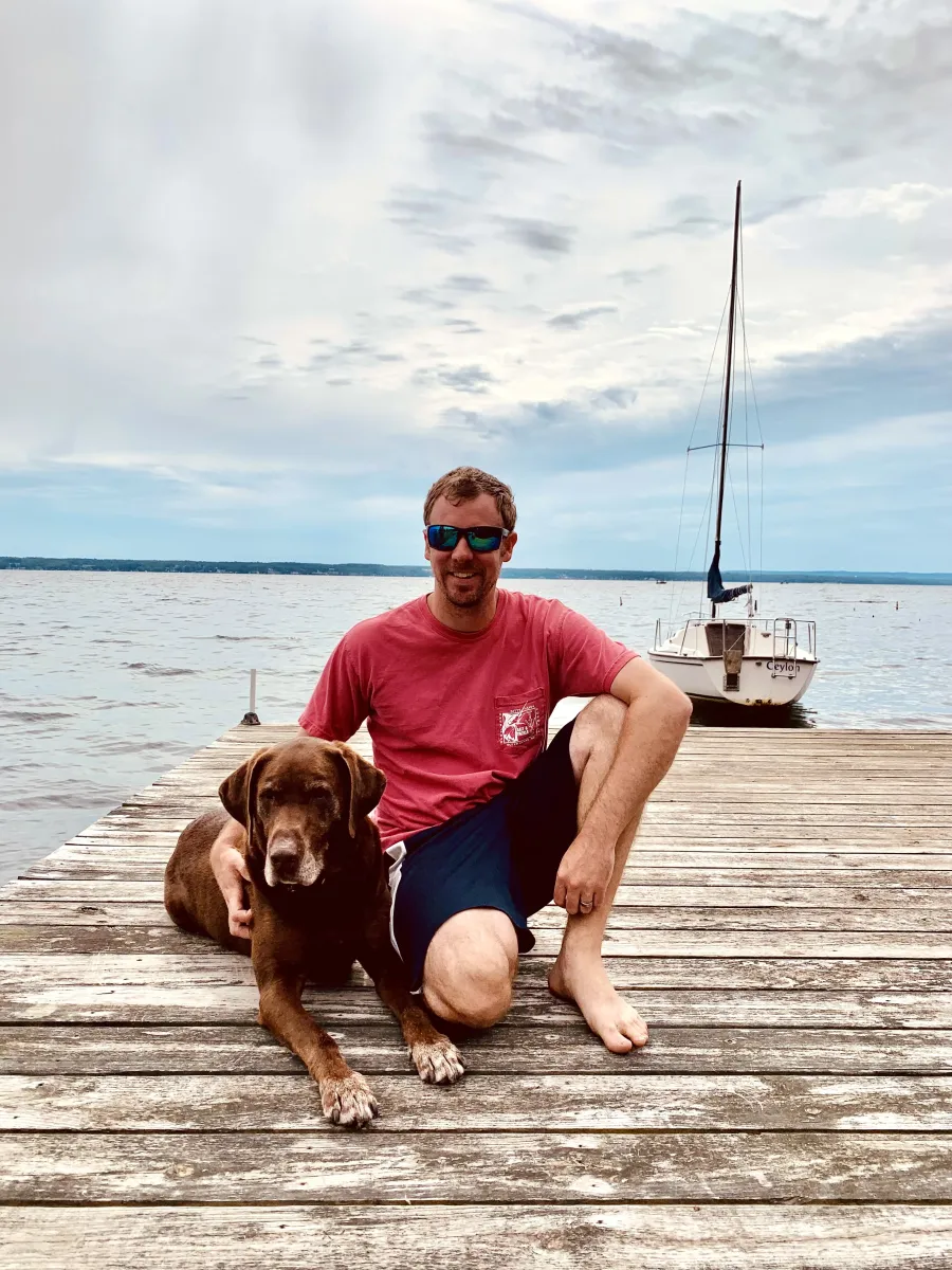 Ryan Denny sitting on a lake dock with Byron, his chocolate Labrador retriever