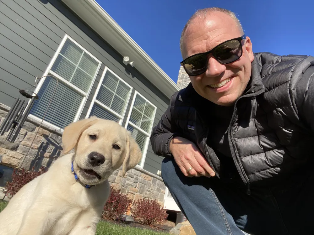 Bill Denny kneeling outside a home with Chloe, his yellow Labrador retriever puppy